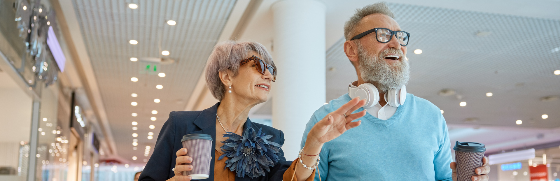 An older couple is walking inside a shopping mall, holding coffee cups IFB